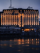 A large, elegant hotel building with multiple windows is illuminated by warm lights, creating reflections on the water in front. The structure's fa&ccedil;ade features classical architectural elements, and an ornate sign is visible on the rooftop. The scene is set during the evening, capturing the serene ambiance of the area.