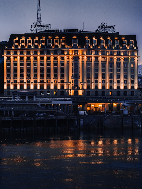 A large, elegant hotel building with multiple windows is illuminated by warm lights, creating reflections on the water in front. The structure's fa&ccedil;ade features classical architectural elements, and an ornate sign is visible on the rooftop. The scene is set during the evening, capturing the serene ambiance of the area.