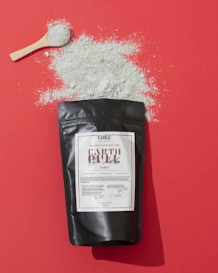 A black package labeled 'EARTH Black Mask Purify' lies on a red surface. There is a wooden spoon on top of the surface with a pile of gray powder scattered around it.
