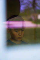 A boy looking out a window with a thoughtful expression, city ruins visible in the background.