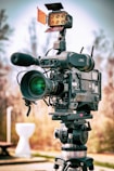 A professional video camera is mounted on a tripod, equipped with various attachments including a microphone and a mounted light with an orange filter. The background is blurred, suggesting an outdoor setting on a clear day.