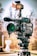 A professional video camera is mounted on a tripod, equipped with various attachments including a microphone and a mounted light with an orange filter. The background is blurred, suggesting an outdoor setting on a clear day.