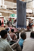 In a busy outdoor market, several people are gathered around a street vendor selling onions and potatoes. The scene is bustling with activity as people sit and stand near a colorful assortment of vegetables. Vehicles and rickshaws are visible in the background, and a cloth hangs prominently in the foreground, partially obscuring the view.