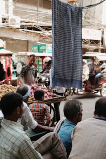 In a busy outdoor market, several people are gathered around a street vendor selling onions and potatoes. The scene is bustling with activity as people sit and stand near a colorful assortment of vegetables. Vehicles and rickshaws are visible in the background, and a cloth hangs prominently in the foreground, partially obscuring the view.