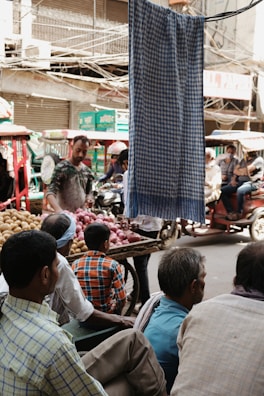 In a busy outdoor market, several people are gathered around a street vendor selling onions and potatoes. The scene is bustling with activity as people sit and stand near a colorful assortment of vegetables. Vehicles and rickshaws are visible in the background, and a cloth hangs prominently in the foreground, partially obscuring the view.