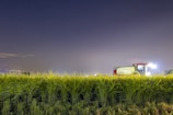 A tractor sits in a field of tall green crops under a clear night sky. Bright lights are visible on the tractor, casting illumination over the crops.