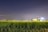 A tractor sits in a field of tall green crops under a clear night sky. Bright lights are visible on the tractor, casting illumination over the crops.