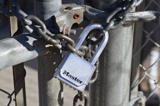 A heavy-duty padlock secures a metal chain wrapped around a galvanized fence post. The lock is silver with visible branding, and the chain is thick and rugged, suggesting security and protection. Sunlight casts shadows, highlighting the metallic textures.