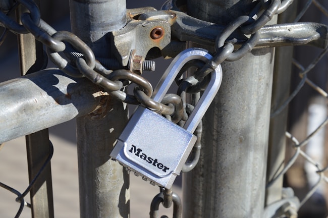 A heavy-duty padlock secures a metal chain wrapped around a galvanized fence post. The lock is silver with visible branding, and the chain is thick and rugged, suggesting security and protection. Sunlight casts shadows, highlighting the metallic textures.