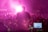 A person stands on stage, silhouetted against bright pink stage lights, facing a large crowd. The person is positioned in front of electronic equipment, including a laptop displaying digital audio software, and they appear to be playing a guitar. The scene is enveloped in vibrant pink and purple lighting, creating an energetic atmosphere.