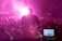 A person stands on stage, silhouetted against bright pink stage lights, facing a large crowd. The person is positioned in front of electronic equipment, including a laptop displaying digital audio software, and they appear to be playing a guitar. The scene is enveloped in vibrant pink and purple lighting, creating an energetic atmosphere.
