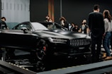 A sleek black luxury car is displayed in an indoor showroom with reflective flooring. Several people are gathered around the car, observing and discussing its features. The environment suggests a high-end car exhibition or launch event.