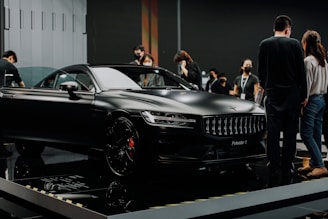 A sleek black luxury car is displayed in an indoor showroom with reflective flooring. Several people are gathered around the car, observing and discussing its features. The environment suggests a high-end car exhibition or launch event.