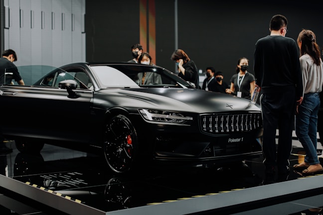 A sleek black luxury car is displayed in an indoor showroom with reflective flooring. Several people are gathered around the car, observing and discussing its features. The environment suggests a high-end car exhibition or launch event.