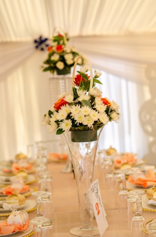 A detailed display table setting inside a white tent, featuring a tall, clear vase filled with white and orange flowers in the center. The table is elegantly set with overturned wine glasses, napkins, and plates, accentuated by a soft peach and cream color palette.