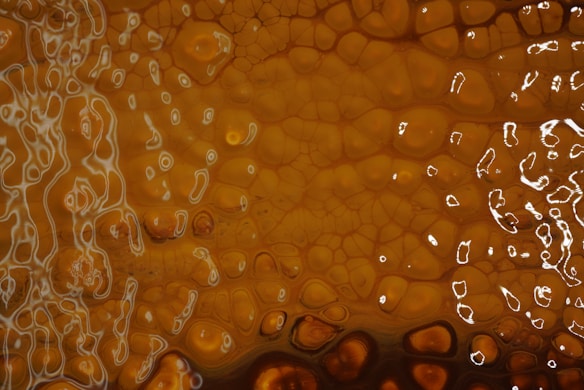 A textured pattern with a rich amber hue, featuring intricate, winding lines resembling veins or cracks. A mix of small, polygonal shapes fills the space, varying in shades of brown and orange. Reflective highlights add a glossy, liquid-like appearance.