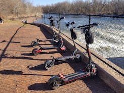Five electric scooters are parked in a row on a reddish gravel path next to a metal fence. Beyond the fence, there is a flowing river with visible rapids. In the background, several people are walking along the path. Bare trees line the riverbank under a clear blue sky.