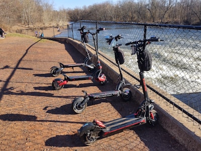 Five electric scooters are parked in a row on a reddish gravel path next to a metal fence. Beyond the fence, there is a flowing river with visible rapids. In the background, several people are walking along the path. Bare trees line the riverbank under a clear blue sky.