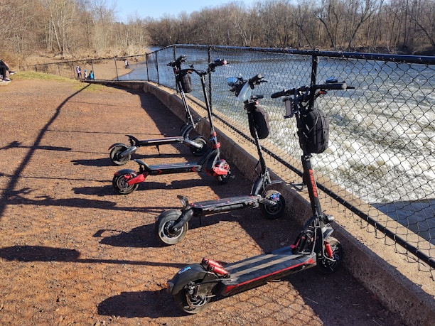 Five electric scooters are parked in a row on a reddish gravel path next to a metal fence. Beyond the fence, there is a flowing river with visible rapids. In the background, several people are walking along the path. Bare trees line the riverbank under a clear blue sky.