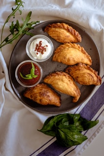 A dark plate features five golden-brown pastries arranged in a semicircle. Next to the pastries are two small bowls, one filled with a white creamy sauce topped with herbs, the other containing a red sauce garnished with fresh mint leaves. Green arugula leaves are placed around the plate, and there is a bunch of basil in the foreground on a white and purple striped cloth.