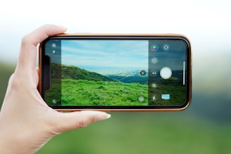 A hand is holding a smartphone horizontally, capturing a photo of a verdant landscape with rolling hills and a partly cloudy sky. The phone's screen displays the photo being taken, showing vivid green grass and hills under a blue sky.