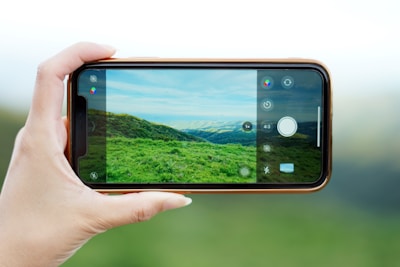A hand is holding a smartphone horizontally, capturing a photo of a verdant landscape with rolling hills and a partly cloudy sky. The phone's screen displays the photo being taken, showing vivid green grass and hills under a blue sky.