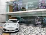 A sleek white sports car is parked on a patterned gray and white tiled pavement. Behind it is a large shop window with the text 'Going Beyond the Ordinary' and floral artwork. The building has modern architecture with glass panels and a wooden accent wall. The reflection in the window shows a city street with additional buildings.