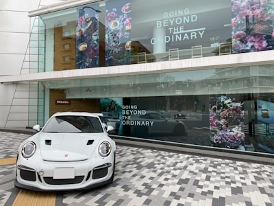 A sleek white sports car is parked on a patterned gray and white tiled pavement. Behind it is a large shop window with the text 'Going Beyond the Ordinary' and floral artwork. The building has modern architecture with glass panels and a wooden accent wall. The reflection in the window shows a city street with additional buildings.