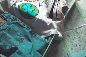 A suitcase is partially packed with a pair of blue jeans, a white T-shirt with a graphic of the Earth, and colorful sneakers. The suitcase is placed on top of a detailed map, suggesting travel plans.