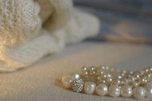 A luxurious composition featuring a strand of lustrous pearls placed on a textured, cream-colored surface. The background shows a soft, knitted fabric, adding a cozy and warm atmosphere to the scene.