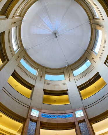 A circular interior with multiple levels of balconies and a large domed ceiling. The architecture features a combination of beige and light brown tones, with modern lighting elements. Blue signage indicates 'Dolby Theatre' at the entrance below the dome.