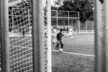 A black and white image capturing an action moment during a soccer game, shot through a netted goalpost. Players are in motion, focusing on dribbling the ball. The field is surrounded by a fence with some trees visible in the background.