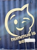 A large, stylized smiley face winking is prominently displayed on a dark blue background. Below the face, there is text saying 'hummus is sexy' in light-colored lettering. A simple white chair sits in the foreground.