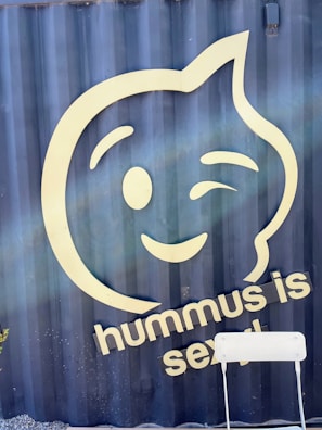 A large, stylized smiley face winking is prominently displayed on a dark blue background. Below the face, there is text saying 'hummus is sexy' in light-colored lettering. A simple white chair sits in the foreground.
