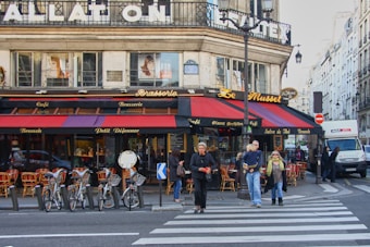 A corner cafe with a classic Parisian facade features a vibrant awning in red and black. Several small tables with red chairs are set outside, and a group of people are crossing the street in front of the cafe. Bicycles are parked along the sidewalk, adding to the urban ambiance. The signage above the windows includes words like 'Brasserie' and 'Cafe', indicating dining options.