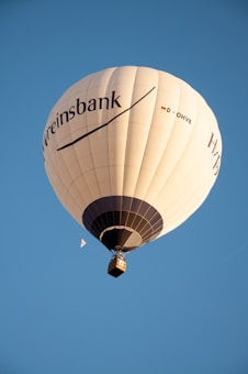 A large white hot air balloon with branding and text ascends into a clear blue sky. The balloon is spherical with vertical panels and a dark stripe near the base. The basket beneath supports a group of people, and there is a small flag attached to the side.