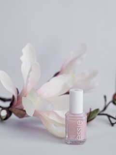 A delicate, pastel pink nail polish bottle is displayed against a soft, light background. The polish bottle is complemented by gentle white and pinkish flowers with green stems, adding a touch of elegance and natural beauty.