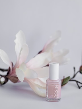A delicate, pastel pink nail polish bottle is displayed against a soft, light background. The polish bottle is complemented by gentle white and pinkish flowers with green stems, adding a touch of elegance and natural beauty.