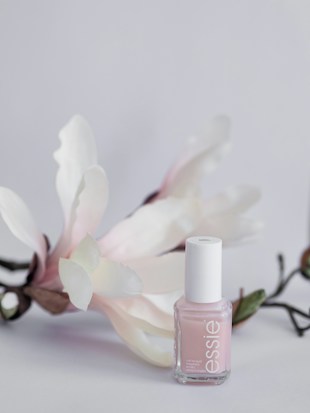 A delicate, pastel pink nail polish bottle is displayed against a soft, light background. The polish bottle is complemented by gentle white and pinkish flowers with green stems, adding a touch of elegance and natural beauty.