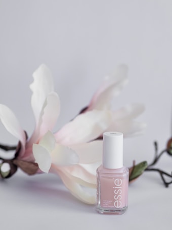 A delicate, pastel pink nail polish bottle is displayed against a soft, light background. The polish bottle is complemented by gentle white and pinkish flowers with green stems, adding a touch of elegance and natural beauty.