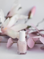A bottle of Essie nail polish is positioned in front of soft pink and white artificial flowers. The background features delicate pastel tones, giving a sense of elegance and subtle beauty.