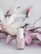 A bottle of Essie nail polish is positioned in front of soft pink and white artificial flowers. The background features delicate pastel tones, giving a sense of elegance and subtle beauty.