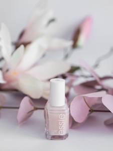 A bottle of Essie nail polish is positioned in front of soft pink and white artificial flowers. The background features delicate pastel tones, giving a sense of elegance and subtle beauty.