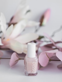 A bottle of Essie nail polish is positioned in front of soft pink and white artificial flowers. The background features delicate pastel tones, giving a sense of elegance and subtle beauty.