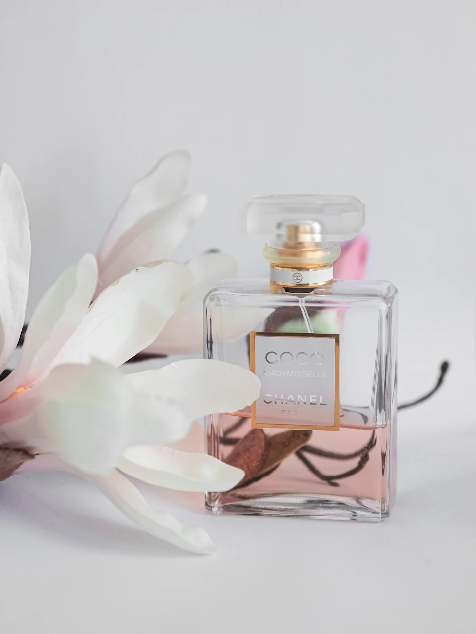 The image features a bottle of Chanel perfume, which is the main subject. The perfume bottle has a clear glass body and a gold cap with a white label that displays "Chanel" on it. The bottle is positioned slightly to the left side of the frame, resting against a backdrop of white flowers.