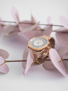 A close-up view of an elegant gold wristwatch with a textured dial, adorned with delicate, pale pink leaves. The watch features a metallic, intricate strap and a shiny bezel that highlights its luxurious design, set against a soft, neutral background.