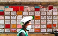 A blurred person wearing a helmet and face mask moves in front of a wall covered with numerous multilingual advertisement flyers. Each flyer is vividly colored with text printed in different fonts and sizes, primarily featuring contact numbers and services.