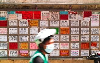 A blurred person wearing a helmet and face mask moves in front of a wall covered with numerous multilingual advertisement flyers. Each flyer is vividly colored with text printed in different fonts and sizes, primarily featuring contact numbers and services.