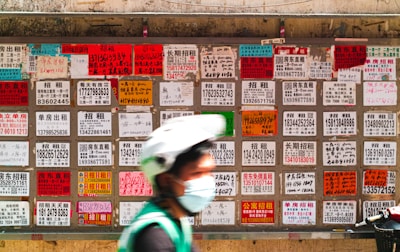 A blurred person wearing a helmet and face mask moves in front of a wall covered with numerous multilingual advertisement flyers. Each flyer is vividly colored with text printed in different fonts and sizes, primarily featuring contact numbers and services.