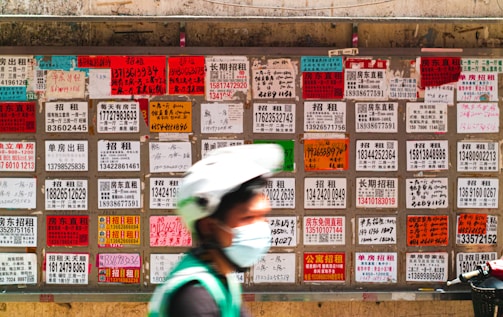 A blurred person wearing a helmet and face mask moves in front of a wall covered with numerous multilingual advertisement flyers. Each flyer is vividly colored with text printed in different fonts and sizes, primarily featuring contact numbers and services.